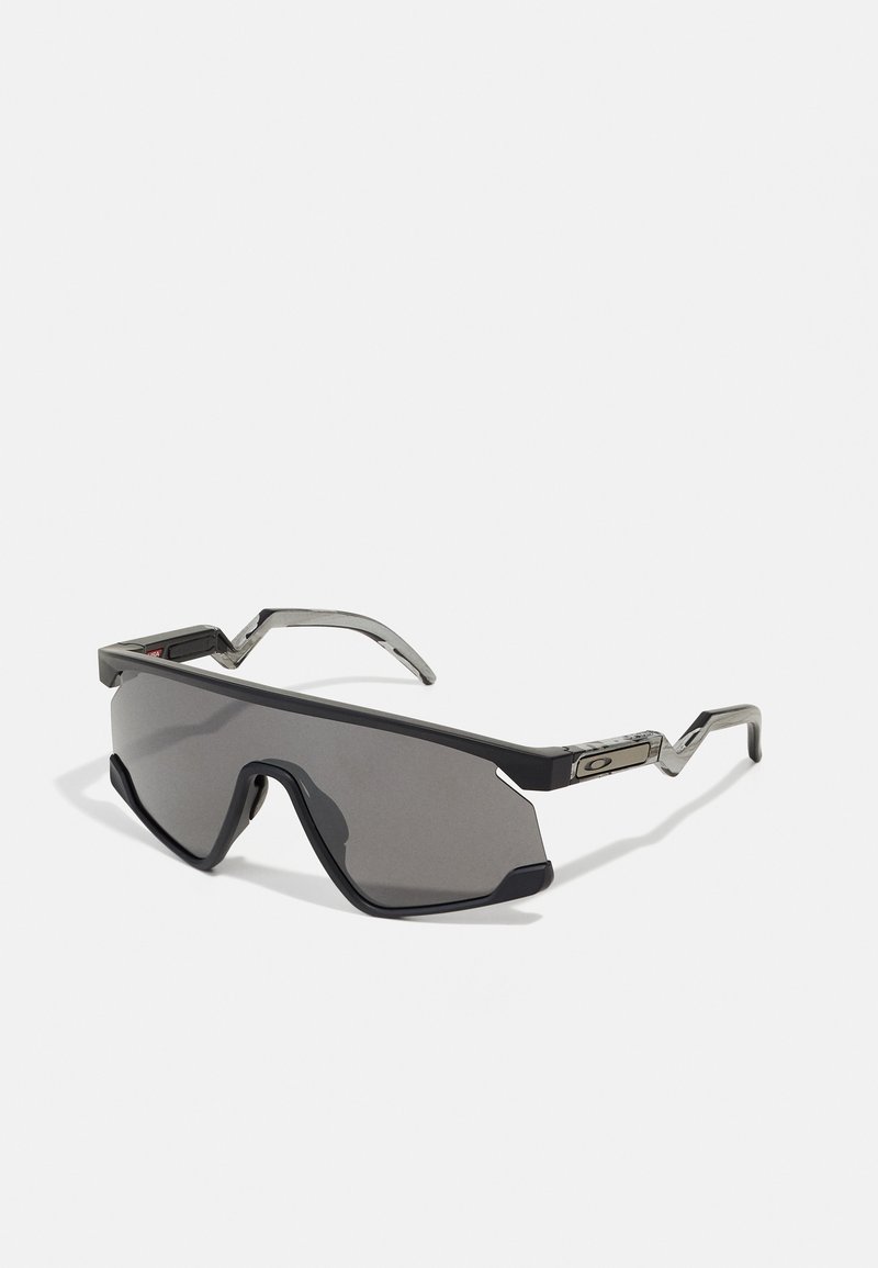 Sonnenbrille mit großem dunklem Glas, schwarzem Kunststoffrahmen und metallischen Akzenten. Abgewinkelte Bügel mit einzigartigem Ausschnitt-Design. Robuste und sportliche Erscheinung.