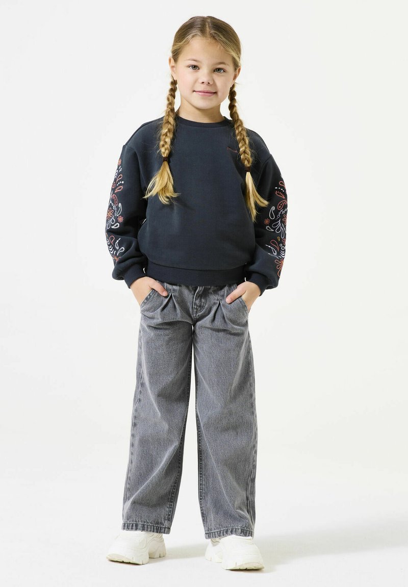 Donkerblauwe sweatshirt met bloemenborduursels op de mouwen, gecombineerd met grijze wijdvallende jeans en witte sneakers. Het model heeft twee gevlochten paardenstaarten.