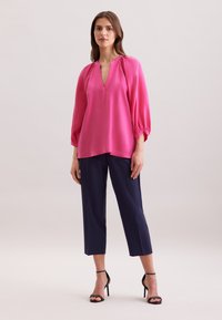 Blusa rosa con scollo a V e dettagli arricciati sulle spalle, abbinata a pantaloni navy a tre quarti e sandali neri con tacco.