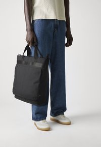Sac fourre-tout en nylon noir avec double poignée, fermeture éclair et devant plat. Présente un design minimaliste avec un accent logo en haut.