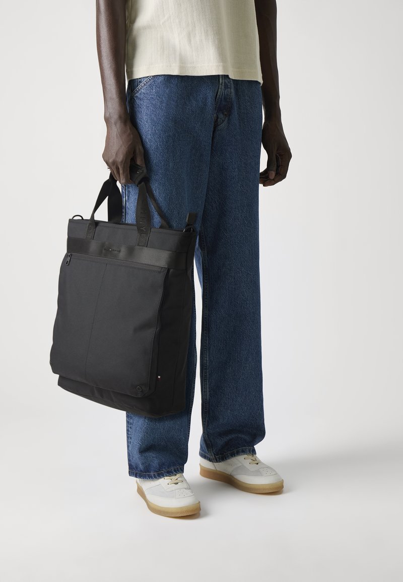 Sac fourre-tout en nylon noir avec double poignée, fermeture éclair et devant plat. Présente un design minimaliste avec un accent logo en haut.