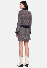 Navy and red tweed jacket with a fitted design, featuring a solid navy collar and waistband. Model wears white heeled boots.