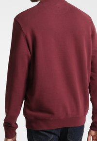 Felpa bordeaux con scollo girocollo e polsini a coste; texture liscia; presenta un design semplice senza motivi o marchi visibili.