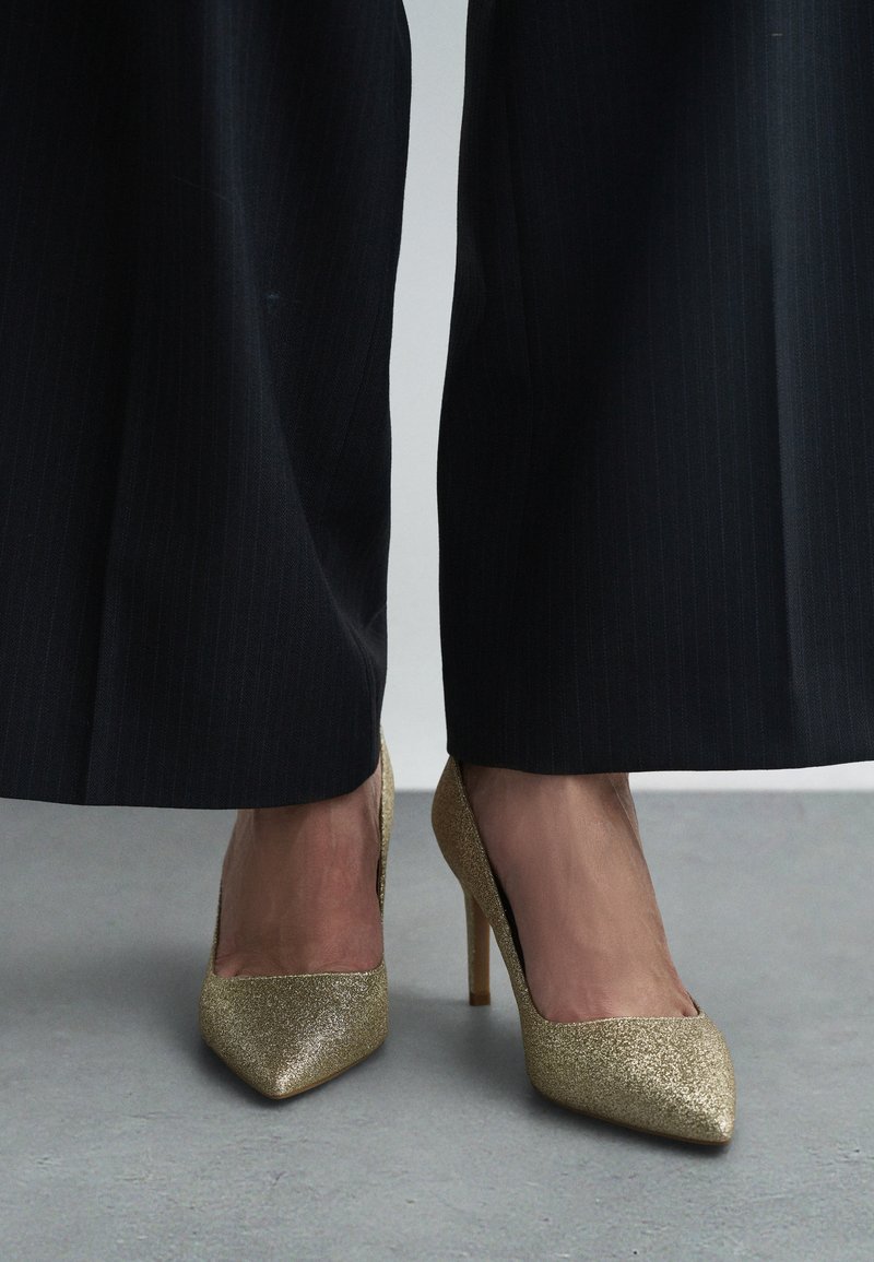 Zlaté výrazné lodičky s špicatou špičkou, v kombinácii s čiernymi širokými nohavicami s prúžkami, ktoré prezentujú elegantný a moderný vzhľad.