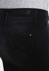 Gros plan de la poche arrière et de la ceinture d'un jean en denim noir porté avec une chemise grise, montrant les passants de ceinture et un patch en cuir marqué.