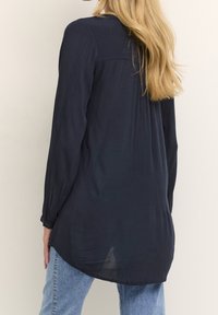 Eine Frau mit langen blonden Haaren, die eine lockere, dunkelblau Langarmbluse und hellblaue Jeans trägt, von hinten vor einem schlichten Hintergrund gezeigt.