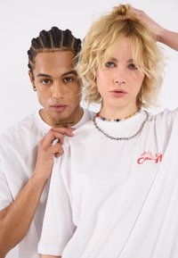 Två individer bär vita t-shirts. Den första skjortan har röd broderad text; den andra har lager av halsband med färgglada pärlor och kedjor.