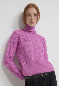 Rosa Rollkragensweater mit Zopfmuster, strukturiertem Finish und geripptem Saum sowie Bündchen, kombiniert mit schwarzen hochtaillierten Hosen.