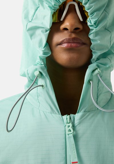 Person wearing a light green hooded jacket with the hood tightened by drawstrings, partially covering the face down to the nose.