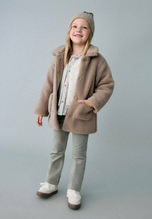 Giacca in pelo finto color beige con colletto, grandi bottoni e tasche frontali. Abbinata a un top bianco con motivo, pantaloni grigi e scarpe bianche con fiocchi.