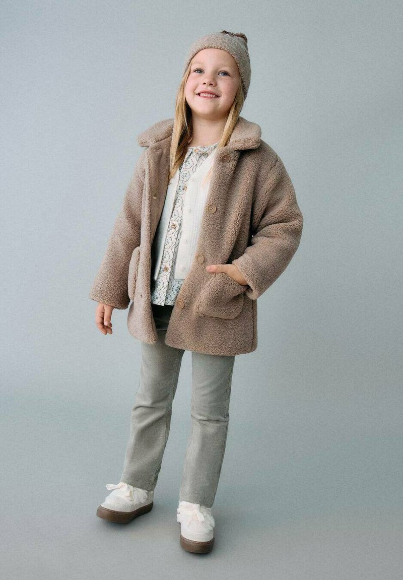 Chaqueta de piel sintética color miel con cuello, botones grandes y bolsillos frontales. Combinada con una blusa blanca de estampado, pantalones grises y zapatos blancos con lazos.