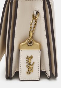 Borsa in pelle color crema con dettagli in marrone scuro, portachiavi dorato e targhetta decorativa con motivo inciso di cavallo e carrozza.