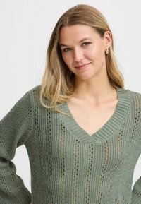 Blond kvinna som bär guldörhängen med ring och en grön stickad V-halsad tröja, ler lätt och står framför en enfärgad ljus bakgrund.