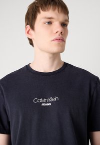 Čierne bavlnené tričko s okrúhlym výstrihom, na prednej strane s bielym logom "Calvin Klein JEANS", hladká textúra a krátke rukávy.