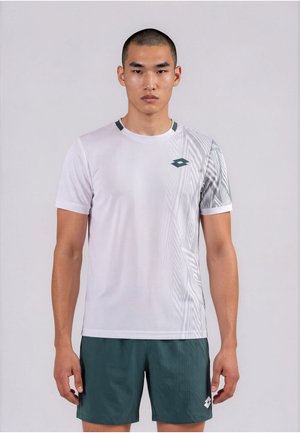 Joven con una camiseta deportiva blanca de manga corta con patrones geométricos y pantalones cortos deportivos de color verde oscuro, de pie con una postura neutral sobre un fondo liso.