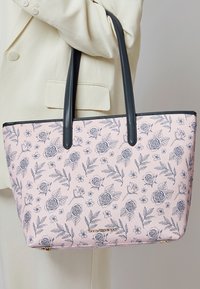 Personne portant un blazer crème tient un sac cabas rose avec un imprimé floral noir et des poignées noires, avec une petite plaque logo dorée.