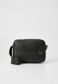 Mala preta retangular tiracolo com alça ajustável, superfície texturizada, nome da marca subtil na frente e um pequeno detalhe em anel prateado.