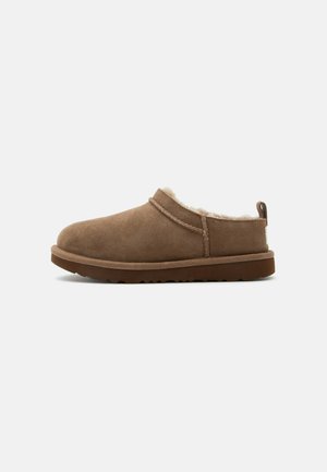 Chausson marron en daim à enfiler avec doublure crème en peluche et semelle en caoutchouc marron, présenté de côté sur un fond clair.