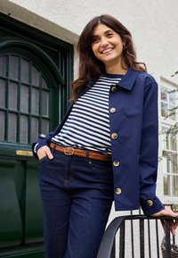 Femme souriante en veste bleu marine et chemise rayée, appuyée sur une rambarde noire devant une porte verte et un bâtiment blanc avec des fenêtres.