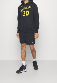 Personne portant un hoodie noir "San Francisco 30", un short noir Nike, des chaussettes blanches et des chaussures de sport noires, debout devant un fond uni.