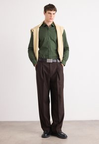 Chemise verte à manches longues, pull beige drapé sur les épaules, pantalon marron, chaussures noires et ceinture grise avec un logo.