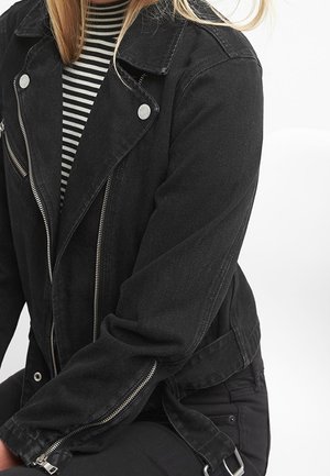 Personne portant une veste biker en denim noir avec des fermetures éclair argentées sur une chemise rayée noire et blanche, assise devant un fond blanc.