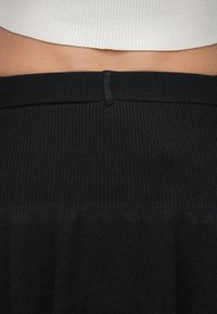 Čierna rebrovaná látka s hladkým textúrou, vybavená priliehavým pásom a skladaným dizajnom na spodnej časti, končiacom v polovici brucha.