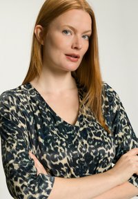 Leopardenmuster-Bluse mit tiefem V-Ausschnitt, dreiviertellangen Ärmeln und einem glatten Stoff in Schwarz, Beige und dezenten blauen Akzenten.