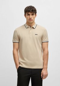 Beige polo skjorte laget av teksturert stoff, med en svart og hvit stripet krage, korte ermer og en subtil logo på brystet.
