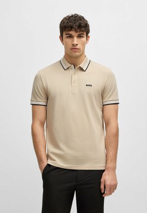 Polo beige realizzato in tessuto testurizzato, con colletto a righe bianche e nere, maniche corte e un logo discreto sul petto.