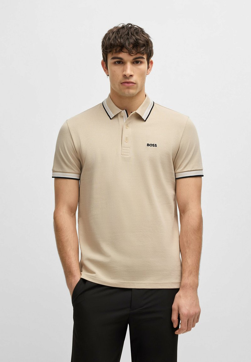 Beige polo skjorte laget av teksturert stoff, med en svart og hvit stripet krage, korte ermer og en subtil logo på brystet.