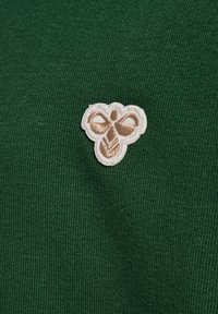 Grönt tyg med ett strukturerat stickmönster, med en liten broderad märke av ett bi i vita och beige nyanser.