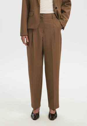 Pantalon marron sur mesure à taille haute porté avec un blazer assorti, un haut blanc et des chaussures noires à bout pointu sur fond blanc.
