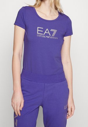 Mujer con una camiseta morada EA7 Emporio Armani y pantalones a juego, de pie frente a un fondo claro y liso.