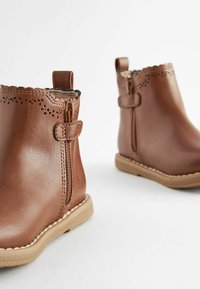 Botas de cuero marrón hasta el tobillo con bordes ondulados, cremalleras laterales y suelas de goma color tostado. Presentan perforaciones decorativas en la parte superior.