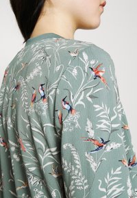 Grön texturerad tyg med färgglad fågel- och bladmönsterbroderi, med intrikata detaljer på axlarna och en ribbad kragsdesign.