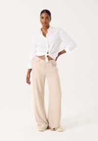 Camisa blanca de botones anudada en la cintura, pantalones beige de pierna larga y zapatos claros. Tela suave con un corte holgado y mangas remangadas.