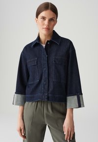 Giacca cropped in denim scuro con due tasche frontali, polsini in denim chiaro a contrasto e chiusura con bottoni. Il modello indossa pantaloni verde oliva.