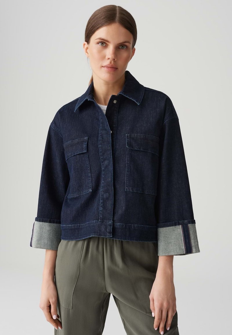 Giacca cropped in denim scuro con due tasche frontali, polsini in denim chiaro a contrasto e chiusura con bottoni. Il modello indossa pantaloni verde oliva.