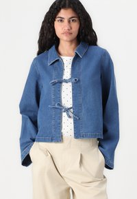 Zilā džinsa jaka ar apkakli, ar mezglu aizdares, brīvā fitā un garām piedurknēm, valkāta virs baltas punktotas blūzes un bēšiem biksēm.