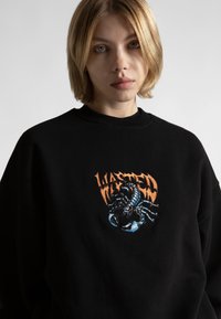 Felpa nera con un vivace design ricamato di uno scorpione e la parola "WASTED" in arancione, su uno sfondo di tessuto texture.