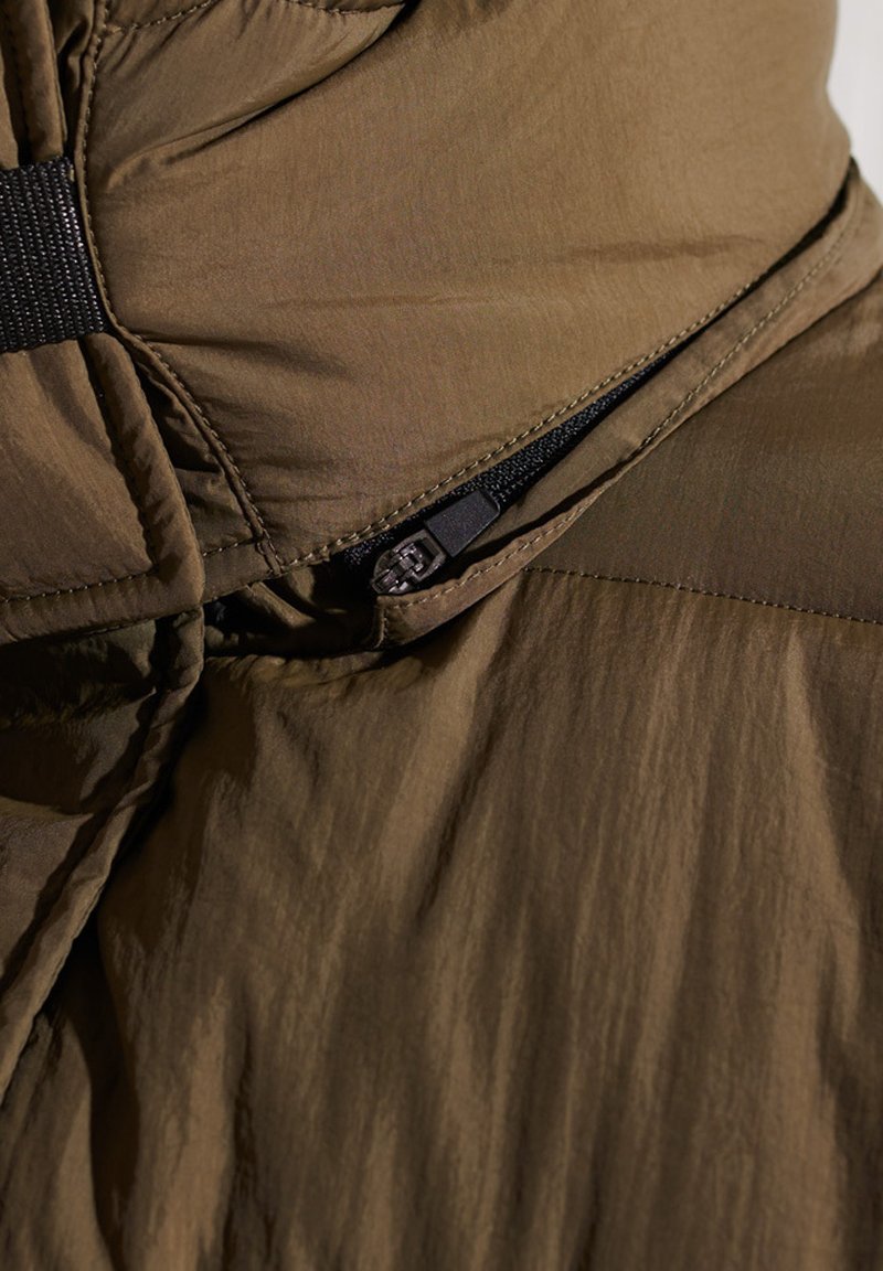 Acercamiento de una cremallera negra parcialmente abierta sobre una tela marrón acolchada, mostrando detalles de costura y textura.