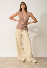 Beżowy, bezrękawnik z głębokim dekoltem w kształcie V, z pięcioma guzikami i przednią kieszonką, zestawiony z kremowymi spodniami szerokimi w pionowe paski.