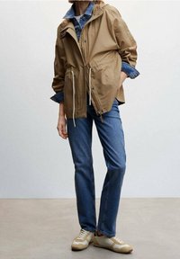 Henkilö, joka on pukeutunut vaaleanruskeaan takkiin farkkutakin päällä, sinisiin farkkuihin ja beigeihin tennareihin seisten yksinkertaista taustaa vasten.