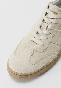 Beige sneakers laget av myk, teksturert semsket skinn, med flate snører, forsterket tå og en gummisåle med lavprofil mønster.