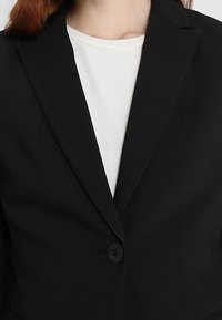 Close-up van een persoon die een zwart gestructureerd blazer draagt over een witte shirt met ronde hals, waarbij de knoop en revers van het blazer zichtbaar zijn.