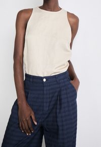 Persoon in een mouwloos beige top die is ingestopt in hoge, marineblauwe, gestreepte broeken, met één hand in de zak en de andere ontspannen.