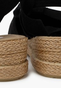 Sandalias espadrilles negras con una media suela de yute trenzado, suela plana y correas de tela ajustables. La textura es suave con un acabado natural.
