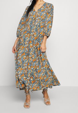 Frau in einem blau, orange und weiß geblümten Midikleid mit dreiviertel Ärmeln und beigen Riemchen-High Heels, stehend vor einem einfarbigen Hintergrund.