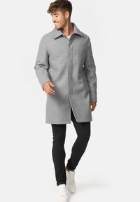 Grå uldfrakke med krave, frontlukning og sidelommer, båret over sorte slim-fit bukser og hvide sneakers. Glat tekstur.